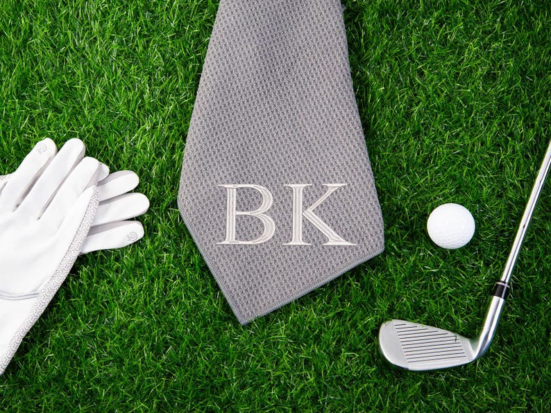 May include: A gray golf towel with the initials "BK" embroidered in white. The towel is lying on green grass next to a white golf ball, a golf club, and a pair of white golf gloves.