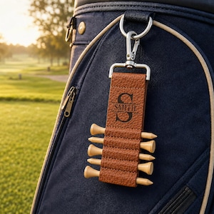 May include: A brown textured leather golf tee holder with a silver metal clip. The leather strap is engraved with a large letter S and the name SMITH. It holds six light-colored wooden golf tees in individual slots, attached to a dark blue golf bag.