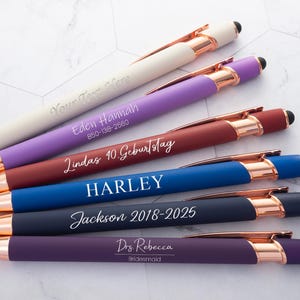 Op de afbeelding: Een assortiment gepersonaliseerde pennen in diverse kleuren, waaronder crème, paars, bordeauxrood, blauw, marineblauw en donkerpaars. Elke pen heeft roségouden accenten en is gegraveerd met aangepaste tekst, zoals namen en data. De pennen hebben een stylus-punt.