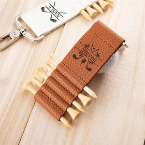 May include: A tan textured leather golf tee holder featuring the name Steven and a golf club graphic. The holder contains five wooden golf tees. A second white golf tee holder with a metal clip and the name Nance is visible nearby.