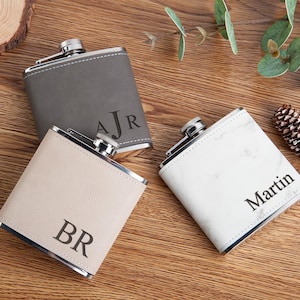 May include: Three personalised hip flasks in grey, beige, and white with silver accents. The grey flask has "AJR", the beige flask has "BR", and the white flask has "Martin" printed on them. The flasks are on a wooden surface.