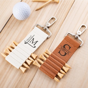 Op de afbeelding: Twee gepersonaliseerde golf tee houders met zilveren sluitingen. Eén is wit met "Mitchell" en een golfclublogo, de andere is bruin met "Smith" en een monogram. Beide houden houten golf tees vast.