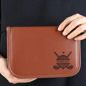 Może przedstawiać: Brązowe skórzane etui na akcesoria golfowe z czarnym paskiem. Etui posiada wzór piłki i kija golfowego z napisem "JORDAN" pośrodku. Etui jest prostokątne z zaokrąglonymi rogami.