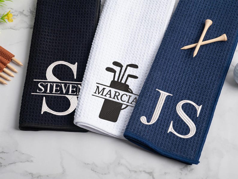 May include: Three golf towels with personalized embroidery. The first towel is black with white embroidery that says "STEVEN". The second towel is white with black embroidery that says "MARCIA" and a golf bag with clubs. The third towel is navy blue with white embroidery that says "JS".