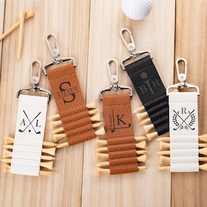 May include: Four rectangular faux leather golf tee holders in white, tan, and black, each featuring a silver metal swivel clasp and personalized engravings. The holders are designed with horizontal slots to secure wooden golf tees in place.