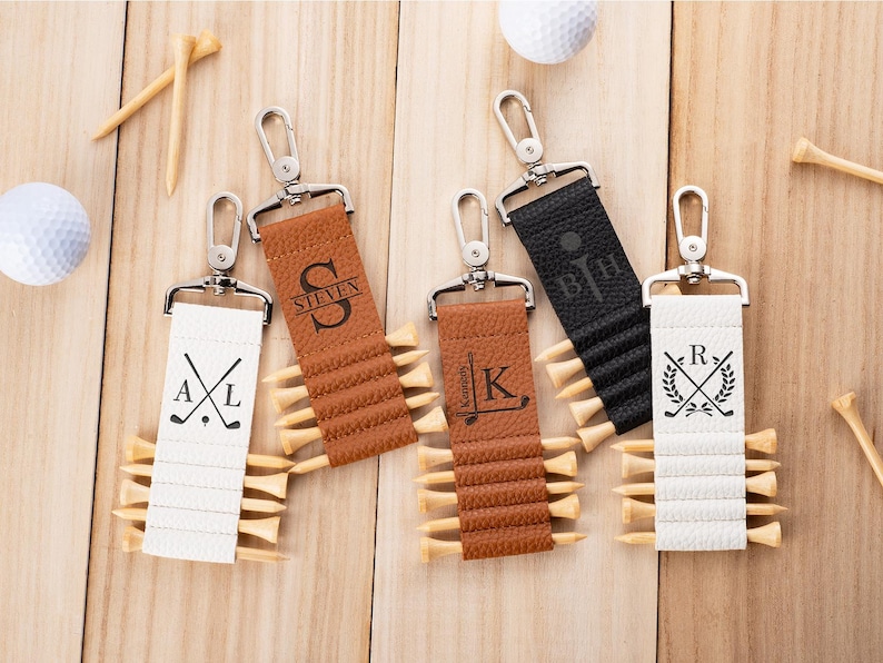 May include: Four rectangular faux leather golf tee holders in white, tan, and black, each featuring a silver metal swivel clasp and personalized engravings. The holders are designed with horizontal slots to secure wooden golf tees in place.