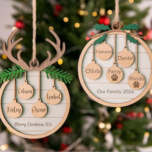 Puede incluir: Dos adornos navideños redondos de madera con listones horizontales claros y blancos. El adorno izquierdo presenta astas marrones y agujas de pino verdes con cuatro círculos colgantes etiquetados como Edison, Isabel, Kaley y Oscar, y Merry Christmas 2026.