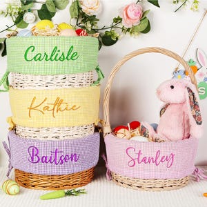 May include: Easter baskets with gingham fabric liners in green, yellow, and purple, each embroidered with a name. A pink basket with a handle holds a plush bunny. Decorative Easter eggs and carrots are scattered around.