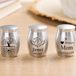 May include: Three small, silver-colored metal urns with rounded bodies and flat lids. The first features a tree engraving with the text Calvin 1936-2024. The second shows angel wings and footprints with James 1948-2024. The third reads In Memory Mom with a heart and 5.12.1977-24.3.2024.