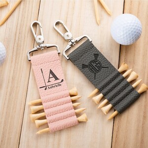 May include: Two textured leather golf tee holders with silver metal swivel clips. One is light pink with the letter A and the name Aubrielle printed above a golf club graphic. The other is dark gray with a circular monogram and golf club graphic.