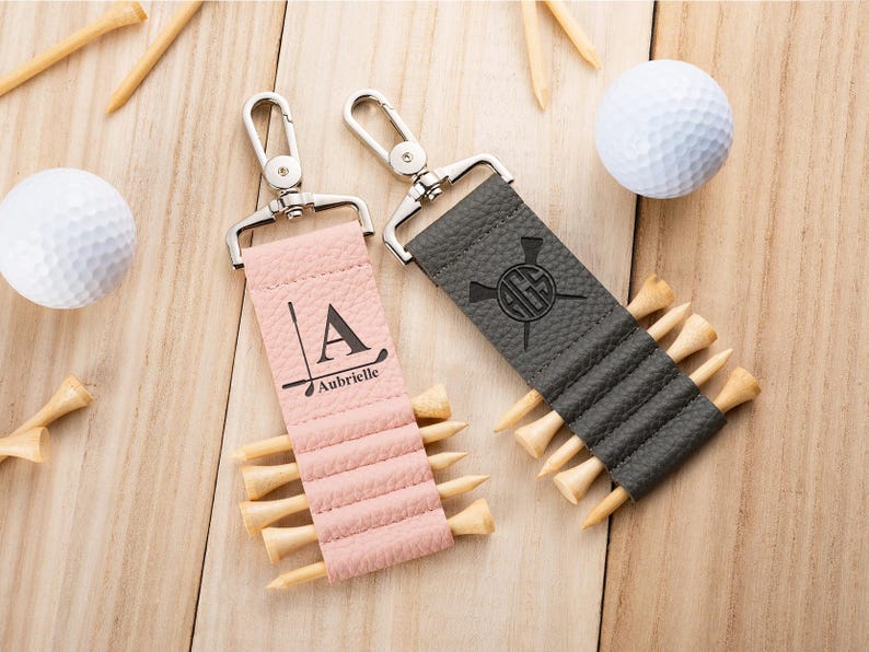 May include: Two textured leather golf tee holders with silver metal swivel clips. One is light pink with the letter A and the name Aubrielle printed above a golf club graphic. The other is dark gray with a circular monogram and golf club graphic.