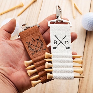 May include: Two textured leather golf tee holders with silver metal clips. One is brown with a black engraved laurel wreath and golf club design with the letter A. The other is white with a black engraved golf club design and letters B and D.