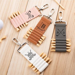 May include: Four rectangular golf tee holders with silver metal lobster clasps. The holders are pink, tan, brown, and white, each featuring custom black text and graphics. They are shown with wooden golf tees inserted and white golf balls nearby.