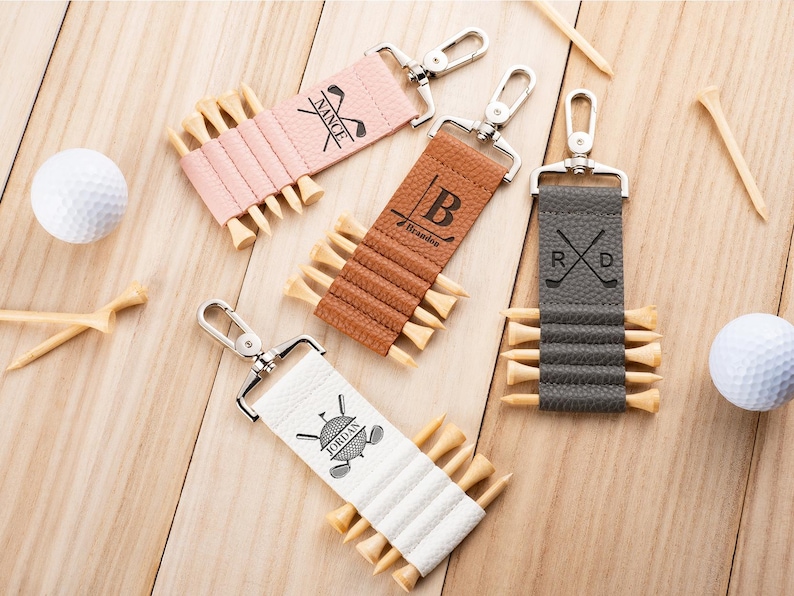 May include: Four rectangular golf tee holders with silver metal lobster clasps. The holders are pink, tan, brown, and white, each featuring custom black text and graphics. They are shown with wooden golf tees inserted and white golf balls nearby.