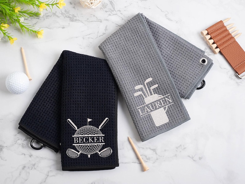 May include: Two personalized golf towels, one gray and one black, with embroidered golf-themed designs and names "Lauren" and "Becker".