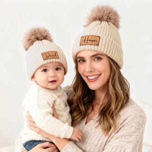 May include: Two cream-colored knit beanies with faux fur pom-poms. One beanie has the name "Daniel" on a leather patch, the other has "Harper". A woman holds a baby wearing the "Daniel" beanie.