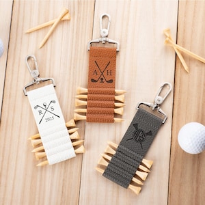 May include: Three rectangular leather keychains in white, tan, and dark gray, each featuring a silver metal swivel clasp and slots holding wooden golf tees. The white keychain displays R, S, and 2025; the tan shows A and H; the gray shows AGS.