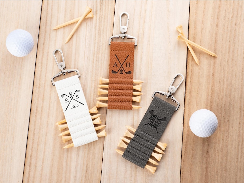 May include: Three rectangular leather keychains in white, tan, and dark gray, each featuring a silver metal swivel clasp and slots holding wooden golf tees. The white keychain displays R, S, and 2025; the tan shows A and H; the gray shows AGS.