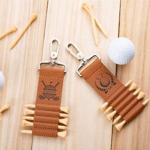 May include: Two tan textured leather golf tee holders with silver metal swivel clips. Each holder features a black engraved golf ball and club graphic with the names Jordan and SR. The holders are filled with light wood golf tees.