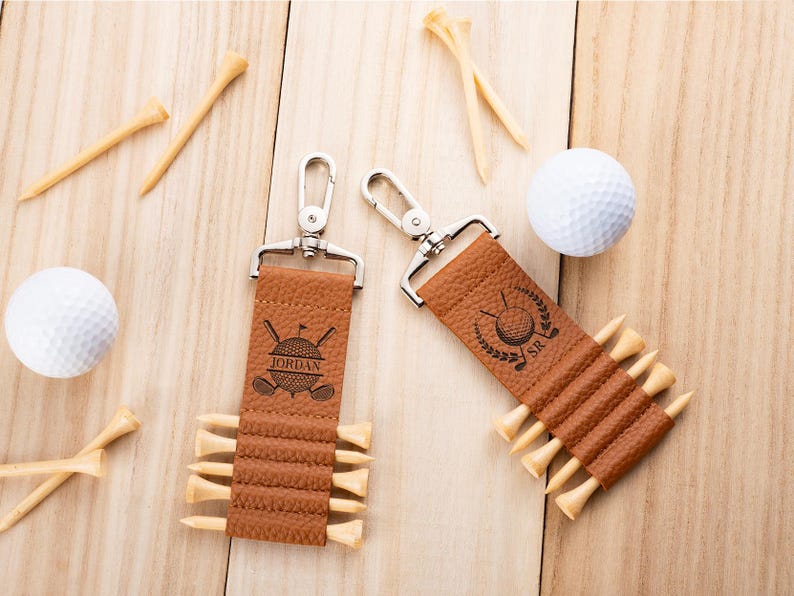 May include: Two tan textured leather golf tee holders with silver metal swivel clips. Each holder features a black engraved golf ball and club graphic with the names Jordan and SR. The holders are filled with light wood golf tees.