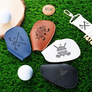 Könnte beinhalten: Eine Sammlung personalisierter Golfaccessoires, darunter Schlägerhauben aus Leder in Blau, Braun, Weiß und Schwarz, mit individuellem Text und Logos. Enthält auch einen Golfball, Tees und einen Schlüsselanhänger-Teehalter, alles auf einer grünen Oberfläche.