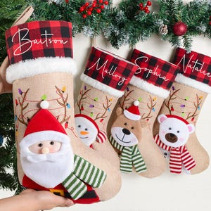 Könnte beinhalten: Weihnachtsstrümpfe mit personalisierten Namen auf rot-schwarz karierten Manschetten. Die Strümpfe sind beige und haben festliche Designs, darunter einen Weihnachtsmann, einen Schneemann, ein Rentier und einen Eisbären.