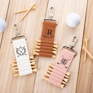 May include: Three textured faux leather golf tee holders in white, tan, and light pink, each featuring a silver metal swivel clasp. The white holder displays a laurel wreath and golf clubs, the tan has an R and Richard, and the pink has HG.