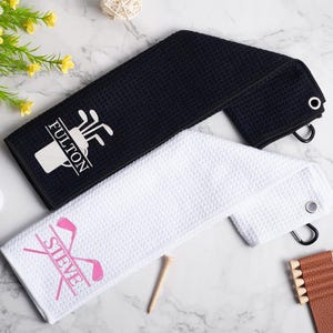 May include: Two white golf towels with black embroidered text. One towel says "FULTON" with a golf bag and clubs design. The other towel says "STEVE" with a pink golf club design.