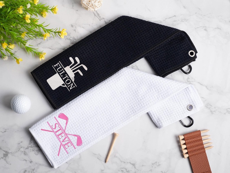 May include: Two white golf towels with black embroidered text. One towel says "FULTON" with a golf bag and clubs design. The other towel says "STEVE" with a pink golf club design.