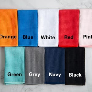 May include: Ten waffle weave kitchen towels in different colors: orange, blue, white, red, pink, green, grey, navy, and black. Each towel has the color name printed on it.