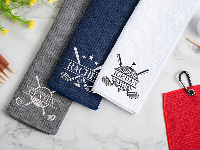 May include: Three golf towels with personalized names, "Rachel", "Jordan", and "Sentry", embroidered in white on a white, navy blue, and gray background. The towels feature a golf ball and golf clubs design.