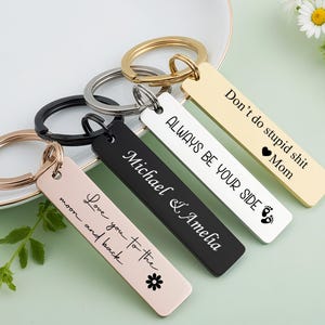 Könnte beinhalten: Eine Auswahl rechteckiger Schlüsselanhänger in Gold, Schwarz, Weiß und Roségold. Jeder Schlüsselanhänger hat einen Metallring und gravierten Text, darunter "Love you to the moon and back" und "Always be your side".