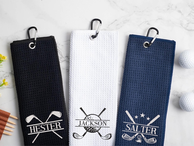 May include: Three personalized golf towels with embroidered names "Hester", "Jackson", and "Salter" and a golf club and ball design. The towels are black, white, and navy blue.