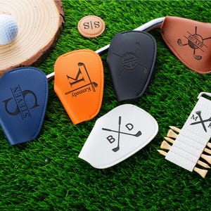 May include: A selection of personalised golf accessories, including club head covers in blue, orange, black, white, and brown. A golf ball and tee holder are also visible. The items are arranged on green artificial turf.