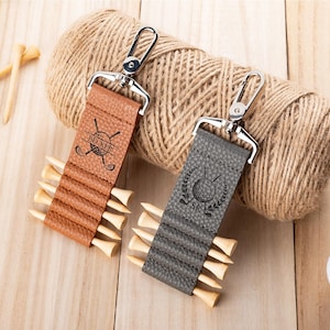 May include: Two textured leather golf tee holders with silver metal swivel clips. The tan holder is engraved with the name BECKER and crossed golf clubs. The gray holder features a golf ball icon, crossed clubs, and the initials SR.