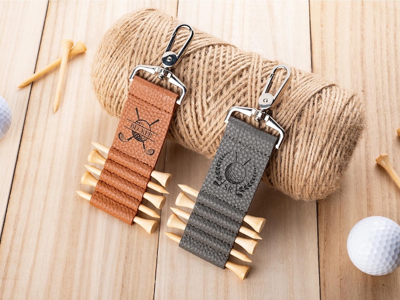 May include: Two textured leather golf tee holders with silver metal swivel clips. The tan holder is engraved with the name BECKER and crossed golf clubs. The gray holder features a golf ball icon, crossed clubs, and the initials SR.
