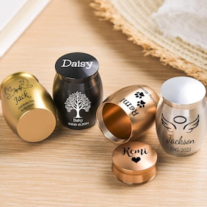 May include: Four small metal cremation urns in gold, black, rose gold, and silver finishes. Each urn features custom engravings including names like Jack, Daisy, Remi, and Jackson, along with dates, paw prints, angel wings, and tree motifs.