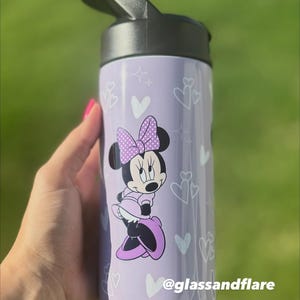 Puede incluir: Una botella de agua lavanda con tapa negra, decorada con corazones y estrellas blancas. Presenta un gr&aacute;fico de Minnie Mouse con un vestido y lazo morados. El texto "@glassandflare" est&aacute; impreso en la botella. Ideal para ni&ntilde;os y fans de Disney.