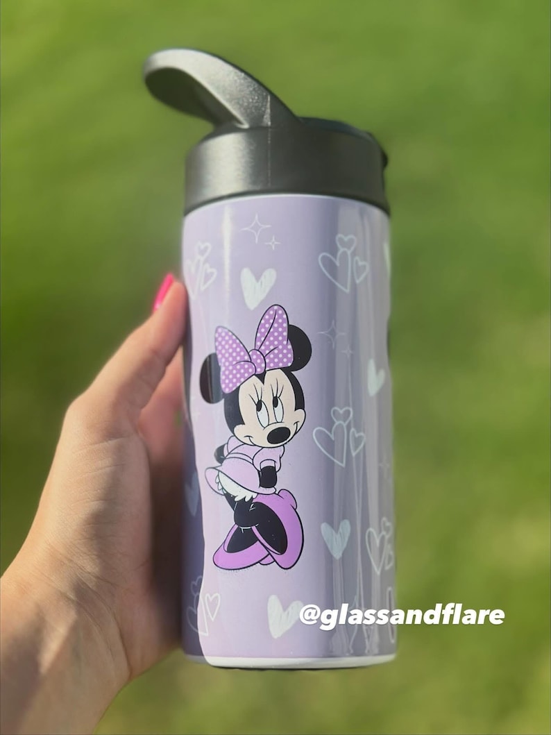 Puede incluir: Una botella de agua lavanda con tapa negra, decorada con corazones y estrellas blancas. Presenta un gr&aacute;fico de Minnie Mouse con un vestido y lazo morados. El texto "@glassandflare" est&aacute; impreso en la botella. Ideal para ni&ntilde;os y fans de Disney.
