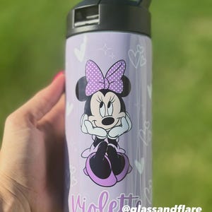 Puede incluir: Una botella de agua morada con Minnie Mouse, un lazo de lunares y el nombre "Violetta". La botella tiene una tapa negra y corazones blancos. Tambi&eacute;n se ve el texto "@glassandflare".