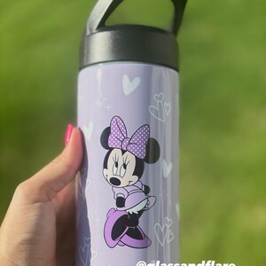 Puede incluir: Una botella de agua morada con un asa negra, decorada con corazones y estrellas blancas. Presenta un dise&ntilde;o de Minnie Mouse con un lazo y un vestido de lunares. El texto "@glassandflare" est&aacute; impreso en la botella.