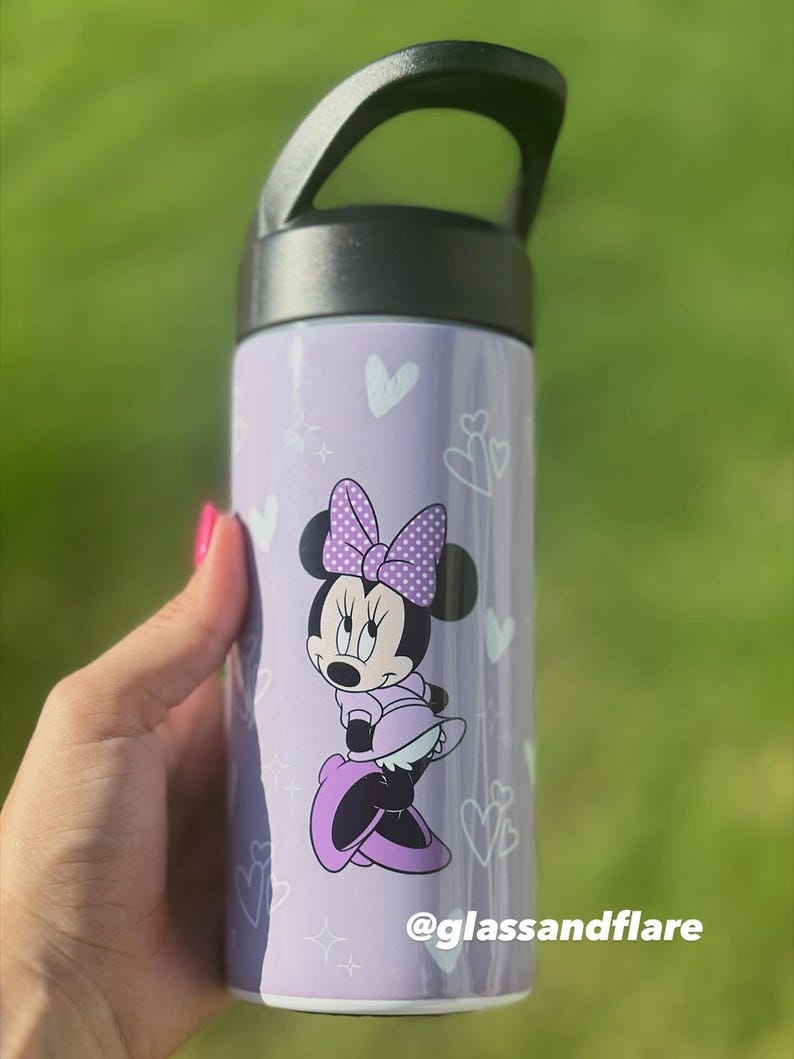 Puede incluir: Una botella de agua morada con un asa negra, decorada con corazones y estrellas blancas. Presenta un dise&ntilde;o de Minnie Mouse con un lazo y un vestido de lunares. El texto "@glassandflare" est&aacute; impreso en la botella.
