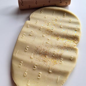 May include: A light beige, textured clay slab featuring embossed numbers and horizontal ridges, dusted with iridescent gold glitter. Positioned above the clay is a cylindrical wooden rolling pin with engraved numbers, used to create the impressions.