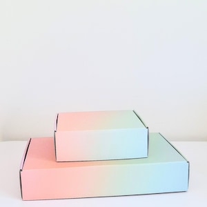 May include: Two rectangular shipping boxes, one atop the other, with a pastel gradient design. The boxes transition from pink to blue-green. The boxes are empty and ready for use.