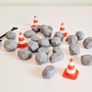 May include: A collection of small, faceted gray stones scattered alongside three miniature orange and white traffic cones and a single black plastic shovel. The stones have a geometric, angular appearance and are arranged in a loose pile.