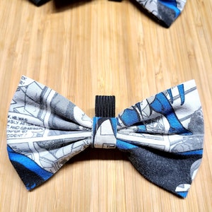 May include: Two bow ties made from comic book pages featuring a superhero. The bow ties are black, white, and blue with a black elastic band.