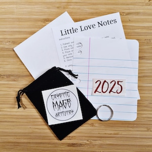 May include: A black velvet pouch with a white circle sticker that says "Cryptic Mage Artistry". The pouch is next to a white sheet of paper with the words "Little Love Notes" and instructions for using the notes. The year 2025 is written in red on the paper.