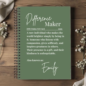 May include: A spiral-bound notebook with a green cover featuring the words "Difference Maker" in elegant script. The definition of "Difference Maker" is printed below, along with the name "Emily". The notebook is set on a wooden surface.