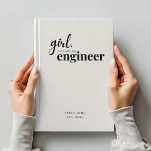 May include: A white journal with the text "girl, you are an engineer" in black script. The journal also includes the name "EMILY JANE" and the year "EST. 2026". The journal is held by a person.