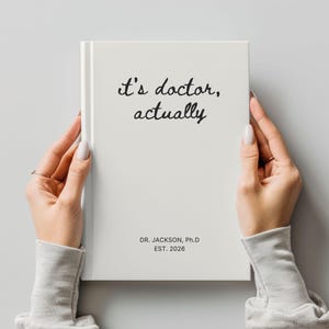 May include: A white hardcover journal with the text "it's doctor, actually" in black script. The journal also includes the text "DR. JACKSON, Ph.D EST. 2026". The journal is held by a person.